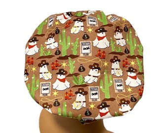 May include: A brown scrub cap featuring a repeating pattern of white ghosts wearing cowboy hats and red bandanas, green cacti, orange cowboy boots, black money bags, yellow stars, and "WANTED" posters with eye masks and dollar amounts.
