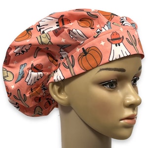 May include: A scrub cap in a dusty pink color featuring a repeating pattern of white ghosts wearing orange cowboy hats, orange pumpkins, green cacti, and blue cowboy boots, accented with small white stars across the fabric surface.