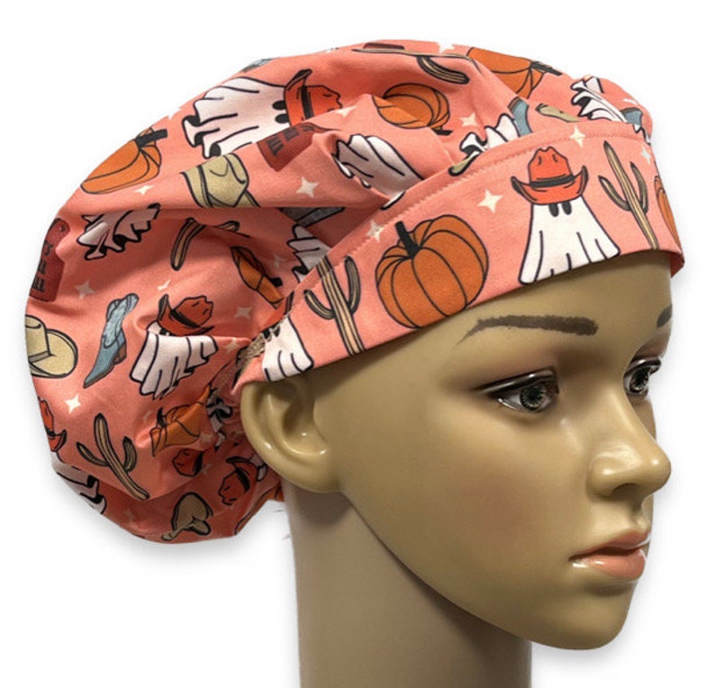 May include: A scrub cap in a dusty pink color featuring a repeating pattern of white ghosts wearing orange cowboy hats, orange pumpkins, green cacti, and blue cowboy boots, accented with small white stars across the fabric surface.