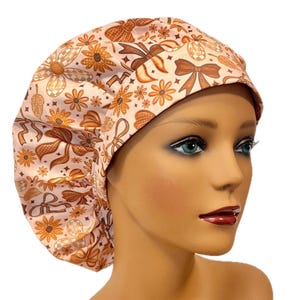 May include: A bouffant scrub cap featuring a light pink background with a repeating pattern of brown bows, orange flowers, and small star shapes. The cap has a wide, structured front band and a gathered back section for a secure, adjustable fit.