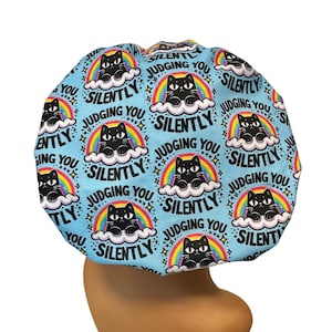 May include: A light blue bonnet with a repeating pattern of black cats on clouds with rainbows. The text "JUDGING YOU SILENTLY" is printed around each cat illustration. The bonnet is designed to cover the head.