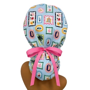 May include: A light blue scrub cap with a repeating pattern of framed coastal images, including seashells, fish, and beach houses. The cap is tied with a bright pink bow. The cap is designed to be worn on the head.