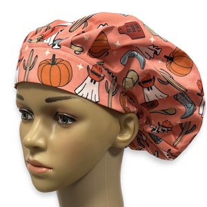 May include: A peach-colored scrub cap featuring a repeating pattern of orange pumpkins, green cacti, white ghosts wearing brown cowboy hats, blue cowboy boots, and red tags with the text IT'S FALL Y'ALL, accented by small white stars.