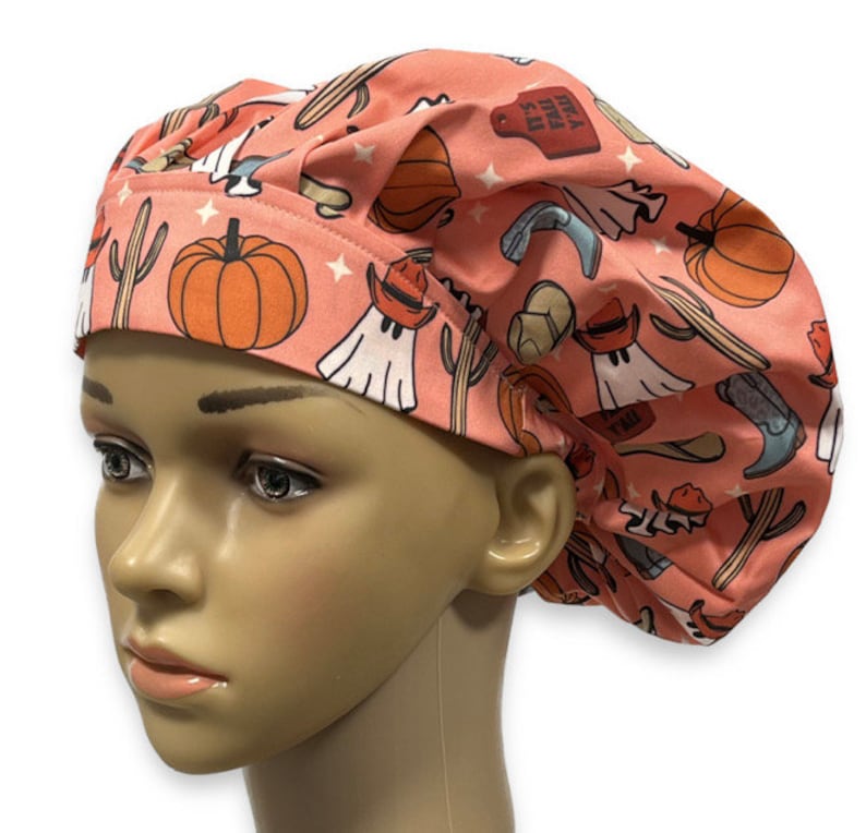 May include: A peach-colored scrub cap featuring a repeating pattern of orange pumpkins, green cacti, white ghosts wearing brown cowboy hats, blue cowboy boots, and red tags with the text IT'S FALL Y'ALL, accented by small white stars.