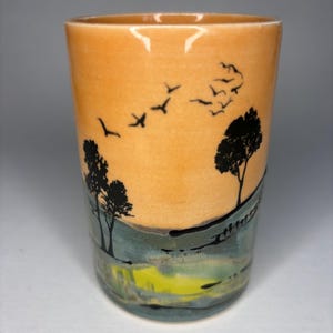 May include: A ceramic tumbler with a landscape design. The tumbler features a sunset scene with black silhouettes of trees and flying birds. The bottom of the tumbler has a green and yellow landscape.