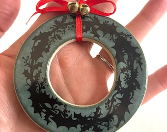 May include: A teal and black Christmas wreath ornament with a holly leaf design. The wreath is adorned with a red ribbon and two small gold bells. This festive decoration is perfect for adding holiday cheer to any space.