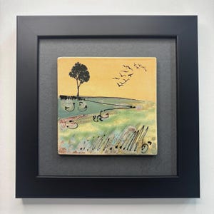 Könnte beinhalten: Gerahmtes Keramikfliesenkunstwerk mit einer Landschaftsszene. Das Kunstwerk zeigt einen Baum, fliegende Vögel und ein Feld mit abstrakten Details in Gelb, Grün und Blau. Der Rahmen ist schwarz.