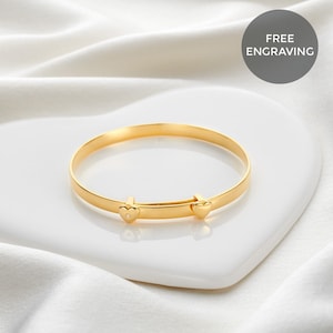 May include: A gold adjustable bangle bracelet with heart-shaped details. The bracelet is presented on a white heart-shaped dish. The words "FREE ENGRAVING" are in a grey circle in the background.