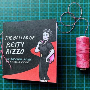 The Ballad of Betty Rizzo, an Abortion Story by Rachelle Meyer, front cover on a cutting mat with pink binding thread