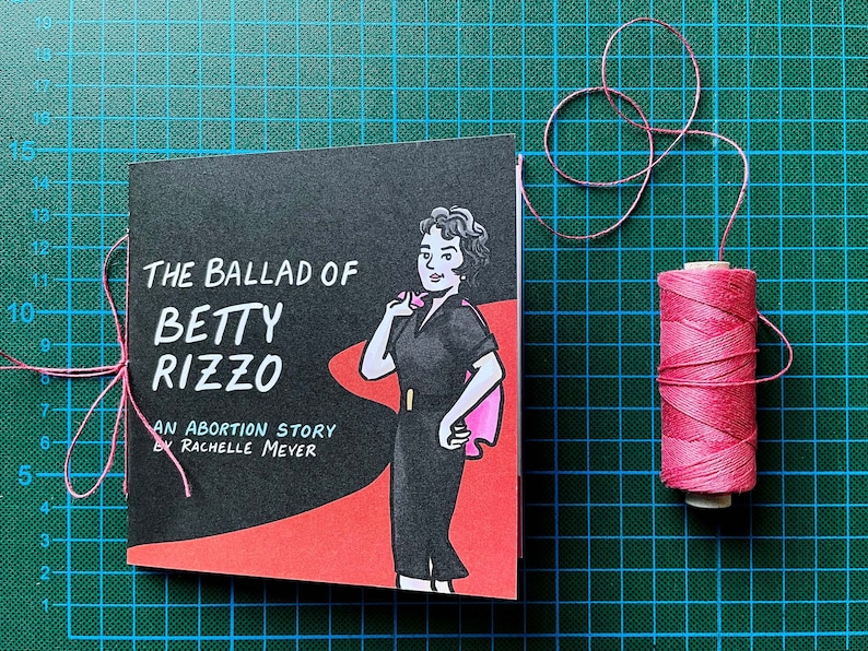 The Ballad of Betty Rizzo, an Abortion Story by Rachelle Meyer, front cover on a cutting mat with pink binding thread