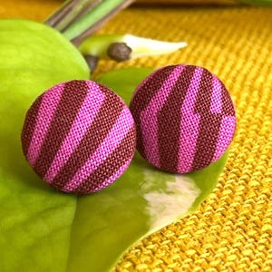 Könnte beinhalten: Zwei runde Ohrringe mit einem gestreiften Muster in Rosa- und Brauntönen liegen auf einem grünen Blatt. Die Ohrringe sind vor einem strukturierten gelben Hintergrund mit grünem Laub im Hintergrund platziert.