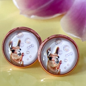 Könnte beinhalten: Ein Paar Ohrstecker aus Roségold mit weißem Hintergrund und einem Cartoon-Lama, das Seifenblasen bläst. Das Lama trägt ein rosafarbenes Stirnband.