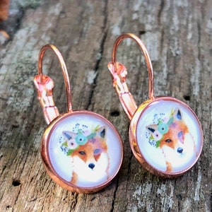 Könnte beinhalten: Ohrringe mit Bügelverschluss in Roségold. Jeder Ohrring zeigt ein rundes, glasbedecktes Bild eines Fuchses mit Blumenkranz. Der Fuchs hat orangefarbenes Fell, ein weißes Gesicht und einen blau-rosa Blumenkranz. Auf einem Holzgrund.