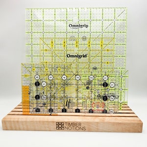 May include: Two yellow Omnigrid rulers with black markings and a smaller orange ruler with black markings. The rulers are on a wooden stand with the words "Timber Notions" engraved on the front.