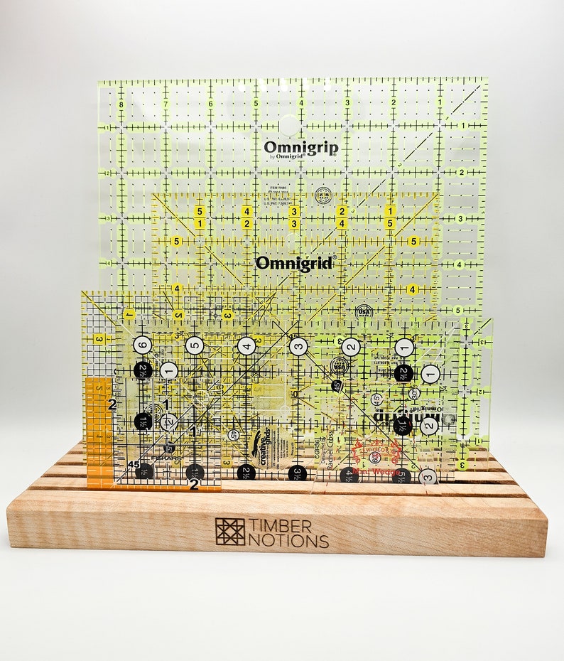 May include: Two yellow Omnigrid rulers with black markings and a smaller orange ruler with black markings. The rulers are on a wooden stand with the words "Timber Notions" engraved on the front.