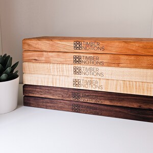 May include: A stack of six wooden cutting boards in various wood tones, including maple, cherry, and walnut. Each board features a laser-engraved logo that reads "Timber Notions".