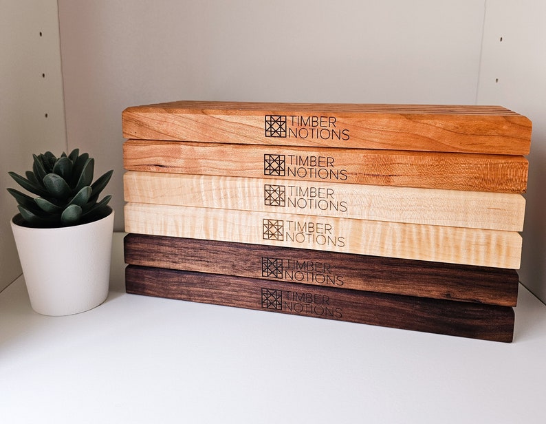 May include: A stack of six wooden cutting boards in various wood tones, including maple, cherry, and walnut. Each board features a laser-engraved logo that reads "Timber Notions".