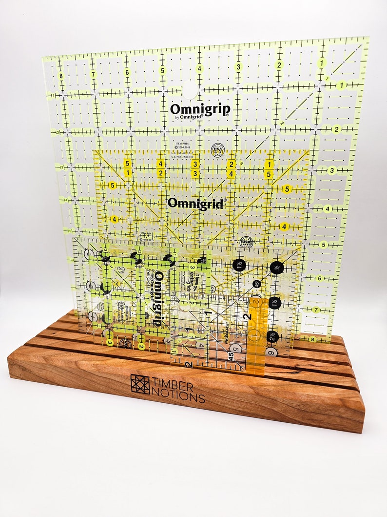 May include: A wooden ruler stand with a yellow and black Omnigrid ruler. The stand is made of cherry wood and has the words "Timber Notions" engraved on the front.