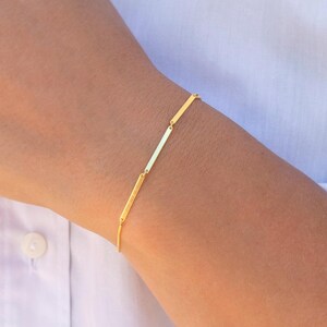 Könnte beinhalten: Ein zartes Goldkettenarmband, das aus einer Reihe kleiner, dünner, rechteckiger Metallstäbe besteht, die in einer durchgehenden Linie miteinander verbunden sind. Das Armband hat eine polierte, reflektierende Oberfläche am Ärmel.