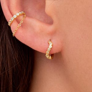 Op de afbeelding: Close-up van een oor versierd met meerdere sieraden. Een kleine oorbel zit bovenaan, een gouden ear cuff met een ketting en een kleine oorring met heldere stenen.