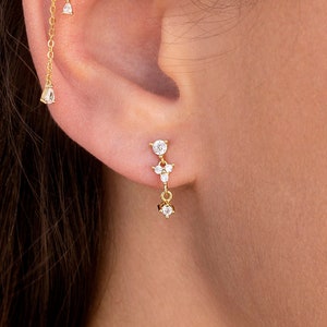 May include: A pair of gold earrings with dangling chains and clear cubic zirconia stones. The earrings are designed with a floral motif and are perfect for adding a touch of sparkle to any outfit.