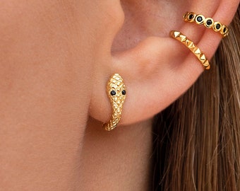 May include: Gold snake ear cuff with black eyes. The snake is curled around the ear and has a textured surface. There are three other gold ear cuffs with black accents.