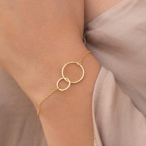 May include: A gold necklace and bracelet with two interlocking circles. The necklace is worn by a person with a light brown shirt.