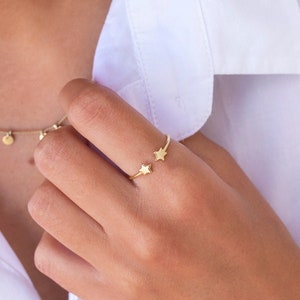 Può includere: Un anello d'oro con due piccoli ciondoli a forma di stella. L'anello è indossato sull'indice di una mano.