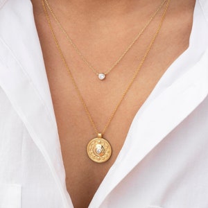 May include: A layered gold necklace with a small round pendant and a larger coin-shaped pendant. The necklace is worn by a person with long dark hair and a white shirt.
