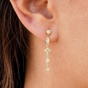Pode incluir: Um conjunto de brincos dourados composto por um pequeno pino com uma pedra pendente de corte marquise e um brinco longo com uma série de pedras transparentes redondas e de corte marquise dispostas numa corrente vertical com um motivo floral.