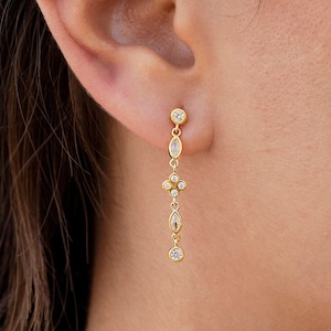 May include: Gold-toned earrings with clear, sparkling gemstones. One earring is a stud with a dangling teardrop shape. The other earring is a long, dangling design with multiple gemstones in various shapes, including round and oval.