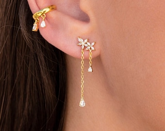 May include: Gold ear cuff with dangling crystal drops and a small crystal flower stud earring. The ear cuff is worn on the upper part of the ear, and the stud earring is worn in the lower lobe.