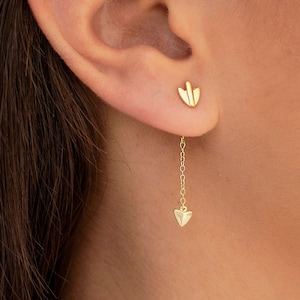 Puede incluir: Un pendiente de aro de oro con un pequeño dije en forma de corazón en la parte inferior y un dije más grande en forma de corazón en la parte superior.