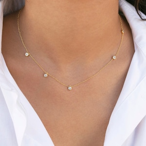 May include: A delicate gold necklace featuring six small, round, clear gemstones evenly spaced along the chain. The necklace is worn against a white collared shirt, creating a simple and elegant look. The necklace is a piece of jewelry.
