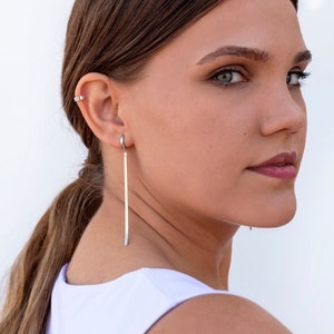 May include: A woman with long brown hair wearing a white vest top and silver earrings. The earrings are long and thin, and they dangle from her earlobes.