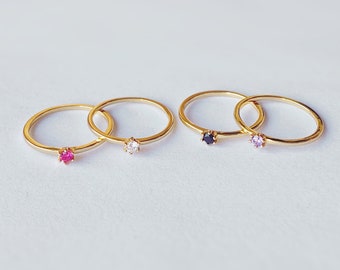 May include: Four gold rings, each with a single gemstone. Two rings have a ruby and a clear stone, while the other two feature a dark blue and a light purple stone. The rings are set against a white background.