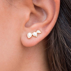 Puede incluir: Un par de pendientes de oro con tres piedras blancas en forma de pera. Los pendientes están diseñados para llevarse en la parte superior de la oreja, con las piedras extendiéndose hacia arriba y hacia afuera.