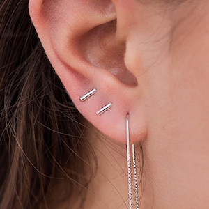 May include: A close-up of a person's ear with two small silver bar earrings and a long silver chain earring hanging down.