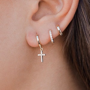 May include: A close-up of a person's ear with multiple gold hoop earrings. The bottom hoop earring has a small cross charm dangling from it.