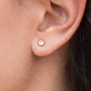 May include: A close-up of a person's ear with a small, clear gemstone stud earring. The earring is set in a gold setting. The person is wearing a delicate gold chain necklace with small gemstones.