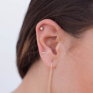 May include: A woman with long brown hair wearing a gold chain necklace and gold earrings. The earrings are a small bar stud and a long chain with a small bar at the end.