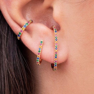 May include: A close-up of an ear adorned with gold-colored jewelry. A small flower-shaped stud earring is at the top. A gold ear cuff and two hoop earrings are embellished with colorful gemstones, creating a rainbow effect.
