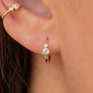 May include: A close-up of an ear displaying gold earrings. The ear has a small hoop earring with three clear stones, a curved ear cuff with three stones, and a dangling stud earring with a clear stone and a teardrop-shaped charm.
