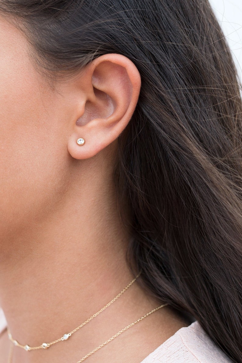 May include: A close-up of a person's ear with a small, clear gemstone stud earring. The earring is set in a gold setting. The person is wearing a delicate gold chain necklace with small gemstones.