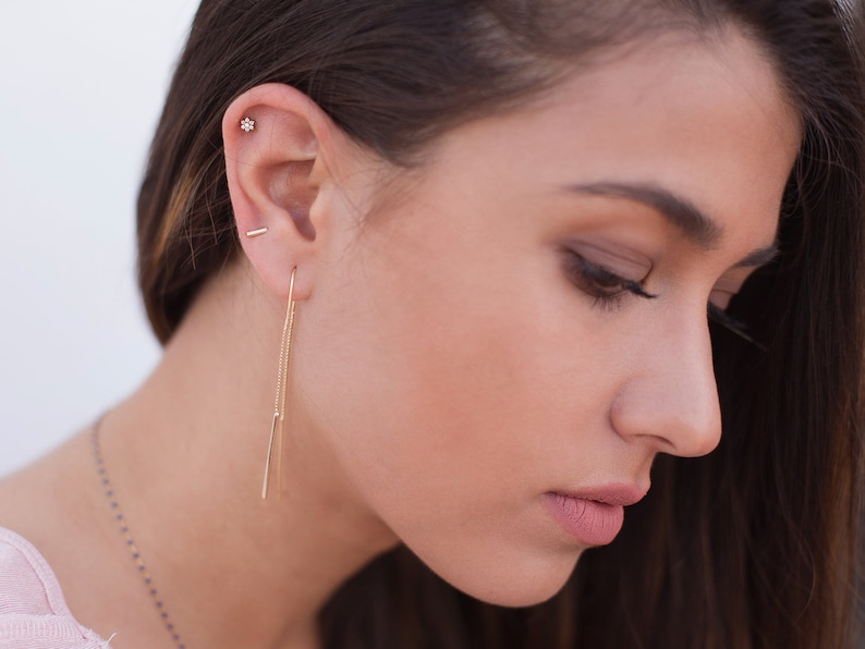 May include: A woman with long brown hair wearing a gold chain necklace and gold earrings. The earrings are a small bar stud and a long chain with a small bar at the end.