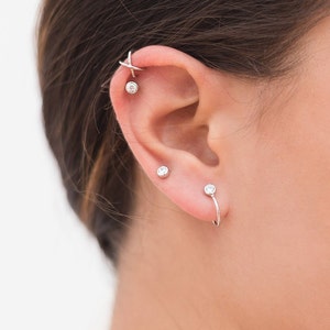 May include: A close-up of a person's ear with multiple silver earrings. The earrings feature small clear gemstones and a unique curved design.