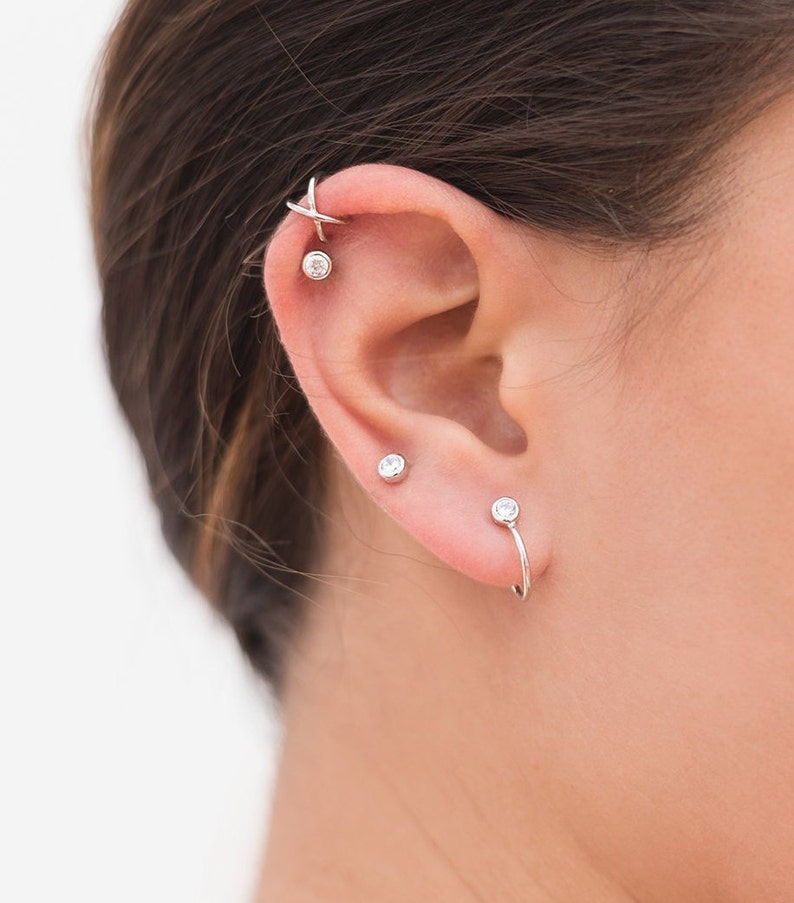 May include: A close-up of a person's ear with multiple silver earrings. The earrings feature small clear gemstones and a unique curved design.