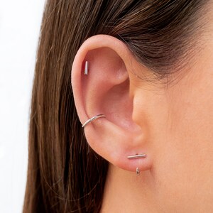 May include: A close-up of a person's ear with three silver earrings. The top earring is a small, straight bar. The middle earring is a thin hoop. The bottom earring is a small, straight bar with a small hoop attached to the bottom.