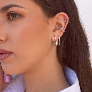 May include: A woman wearing a white shirt with silver hoop earrings in her ear. The earrings are small and have a simple design.