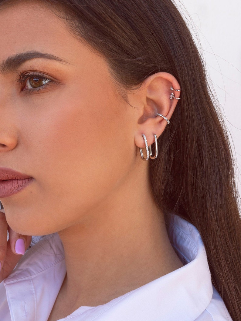 May include: A woman wearing a white shirt with silver hoop earrings in her ear. The earrings are small and have a simple design.